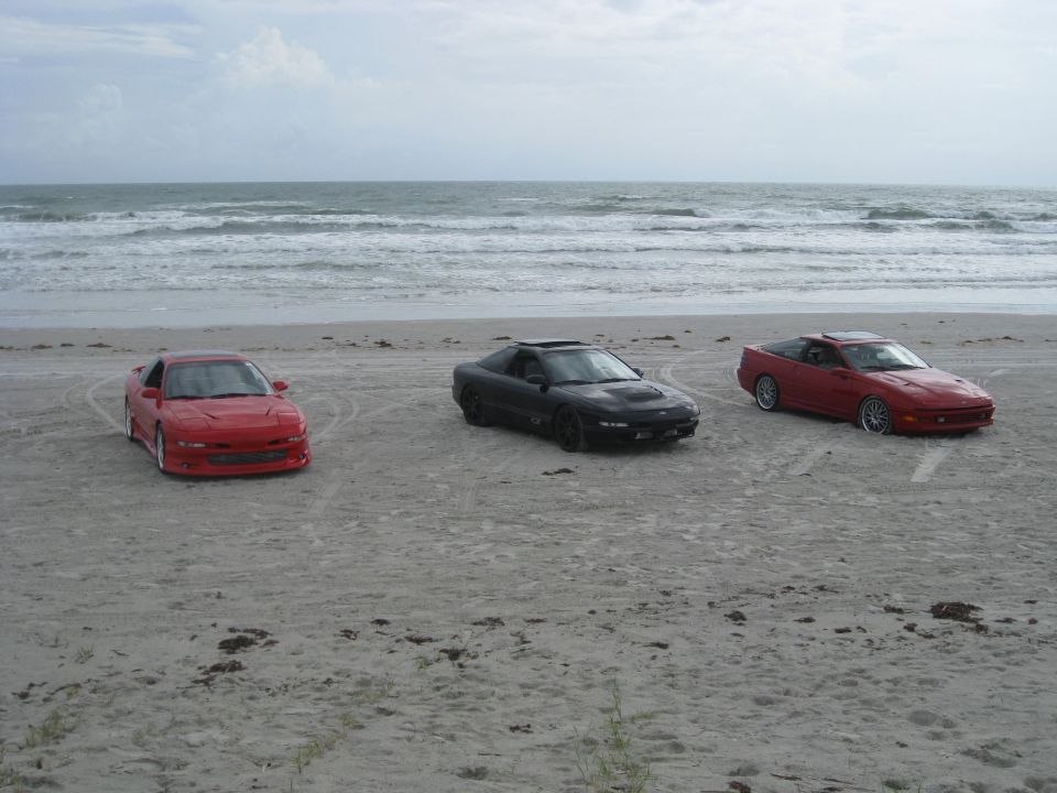 Three sports cars parked on a sandy beach with the ocean in the background. A red car is on the left, a black car in the middle, and another red car on the right