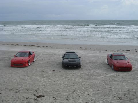 1994 Ford Probe - Three sports cars are parked on the beach near the ocean waves under a cloudy sky.