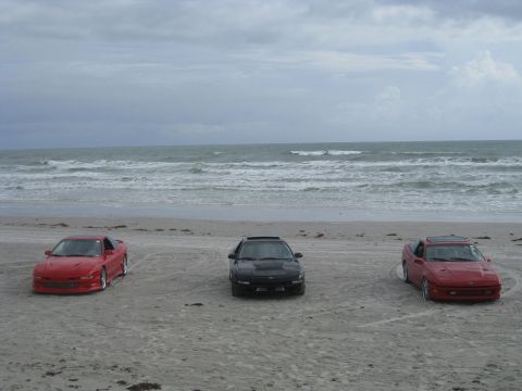 1994 Ford Probe - Three cars are parked on the beach with the ocean waves crashing in the background.
