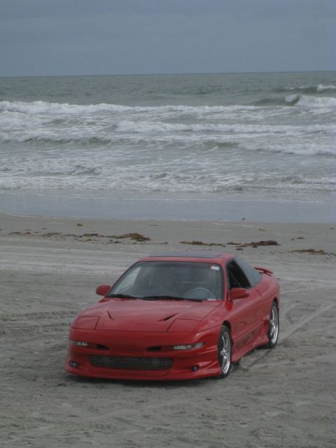1994 Ford Probe - A red coupe with pop-up headlights sits on a beach, with the ocean and sky visible behind it.