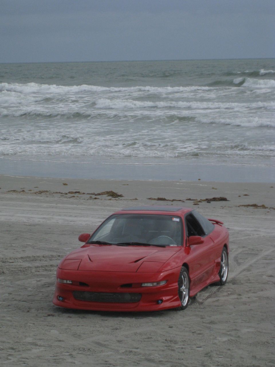Red coupe with pop-up headlights and a sunroof parked on a sandy beach with ocean waves in the background