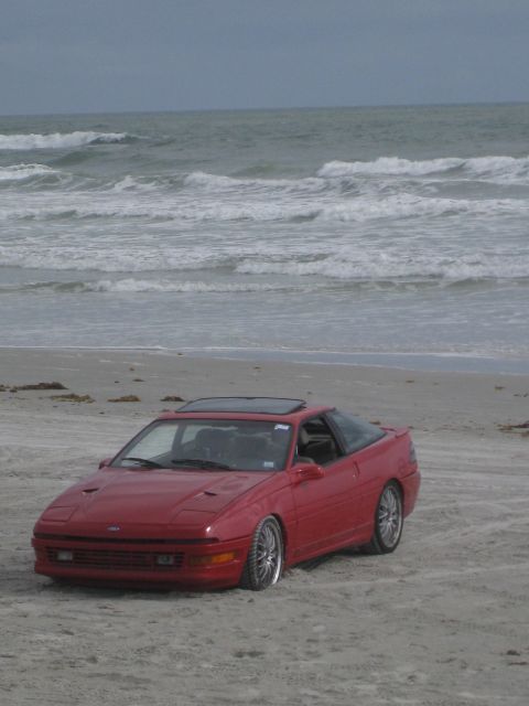 1994 Ford Probe - A red 1994 Ford Probe sits on the sand near the ocean waves.