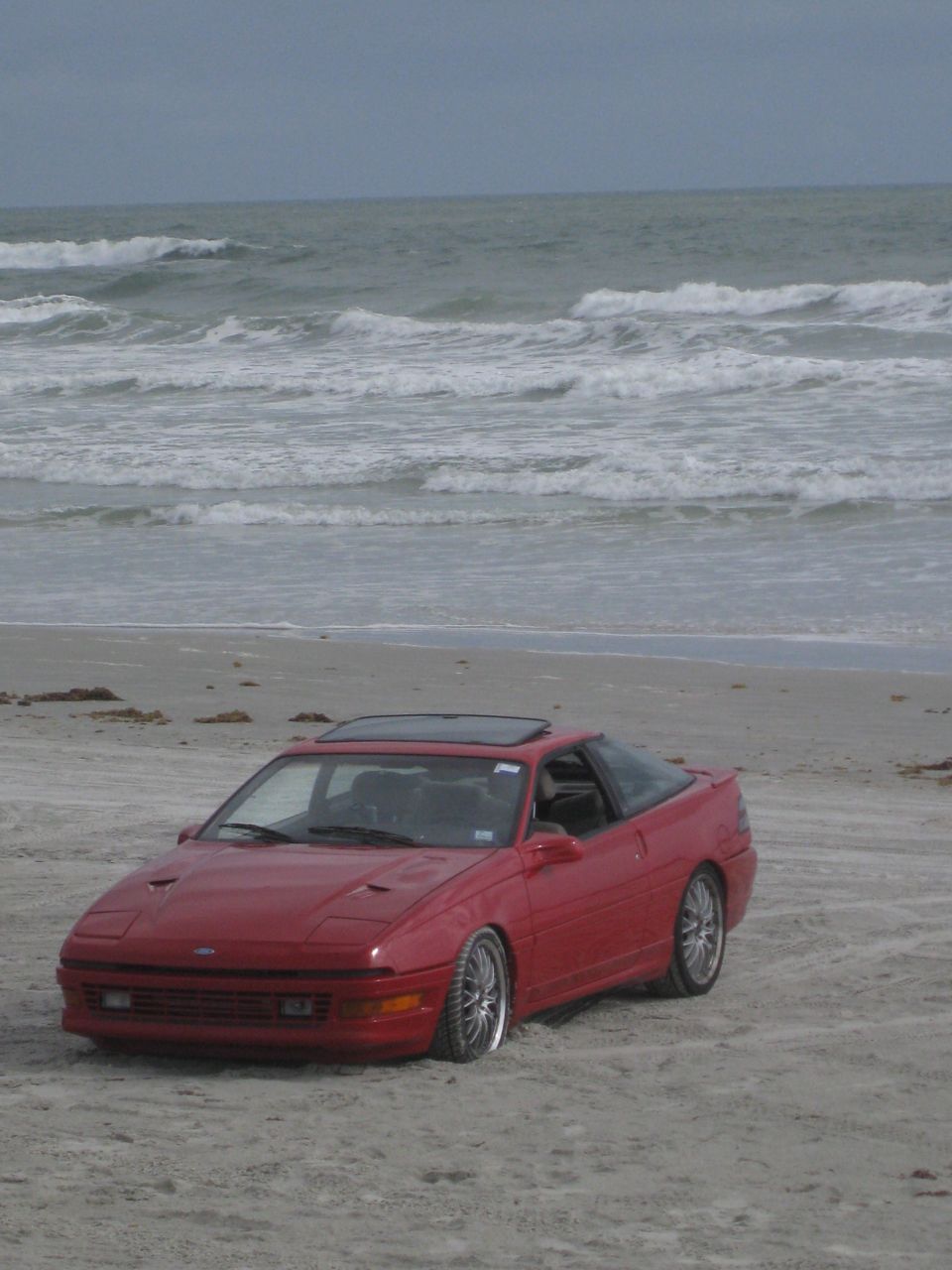 Red 1994 Ford Probe parked on a sandy beach with ocean waves in the background