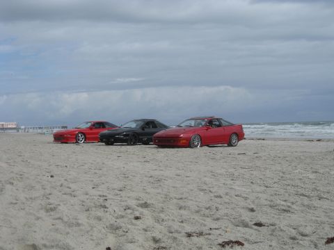 1994 Ford Probe - Three coupes sit on the sand near the ocean waves under a cloudy sky.
