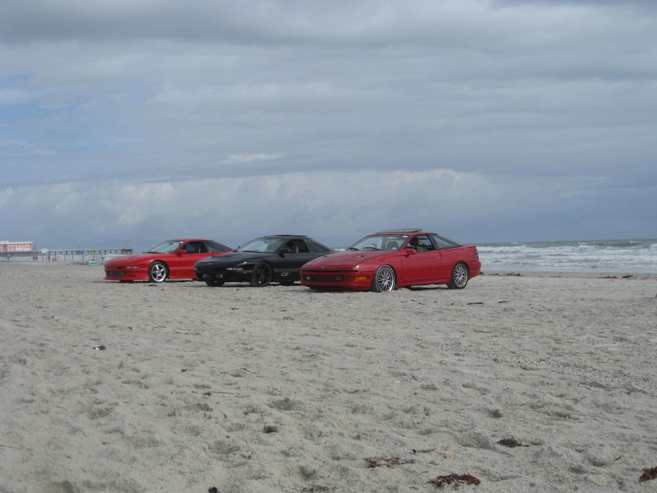 Three sports coupes parked on a sandy beach with the ocean and cloudy sky in the background