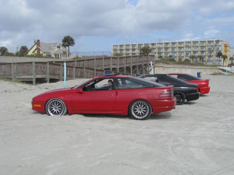 1994 Ford Probe - Three coupes are parked on a sandy beach with a boardwalk and buildings in the background.