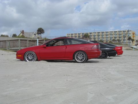 1994 Ford Probe - A red 1994 Ford Probe is parked on a sandy beach, with two other vehicles visible behind it under a cloudy sky.
