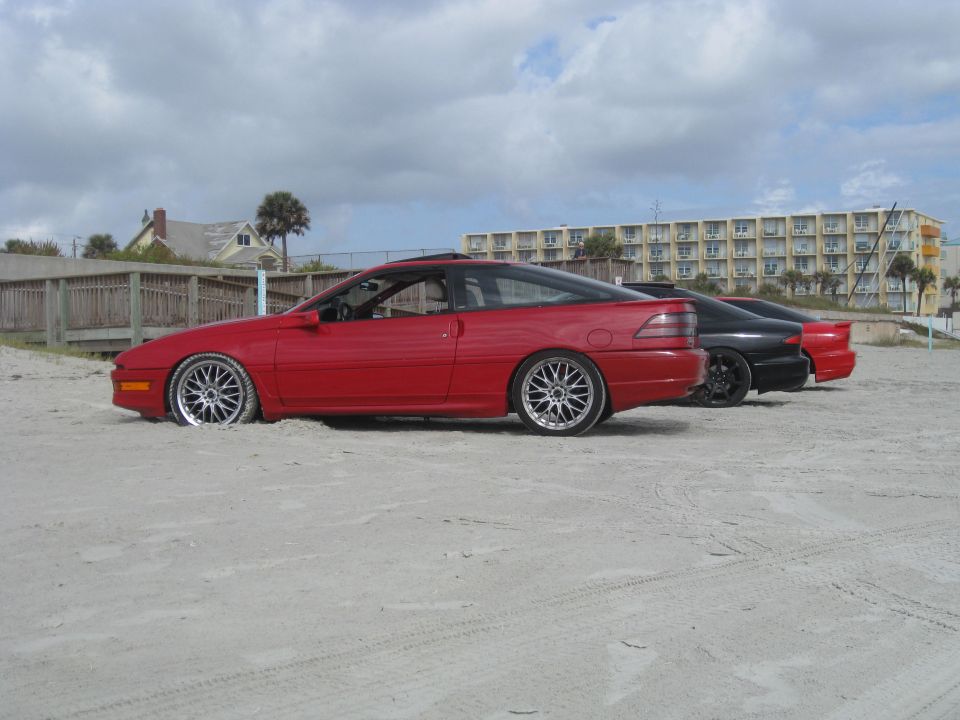 Red 1994 Ford Probe side view on sandy beach with two other cars behind