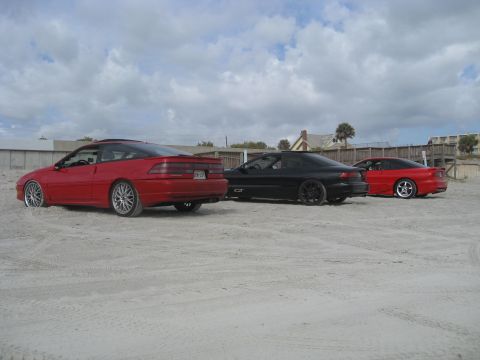 1994 Ford Probe - Three coupes are parked on the sand near buildings under a cloudy sky.