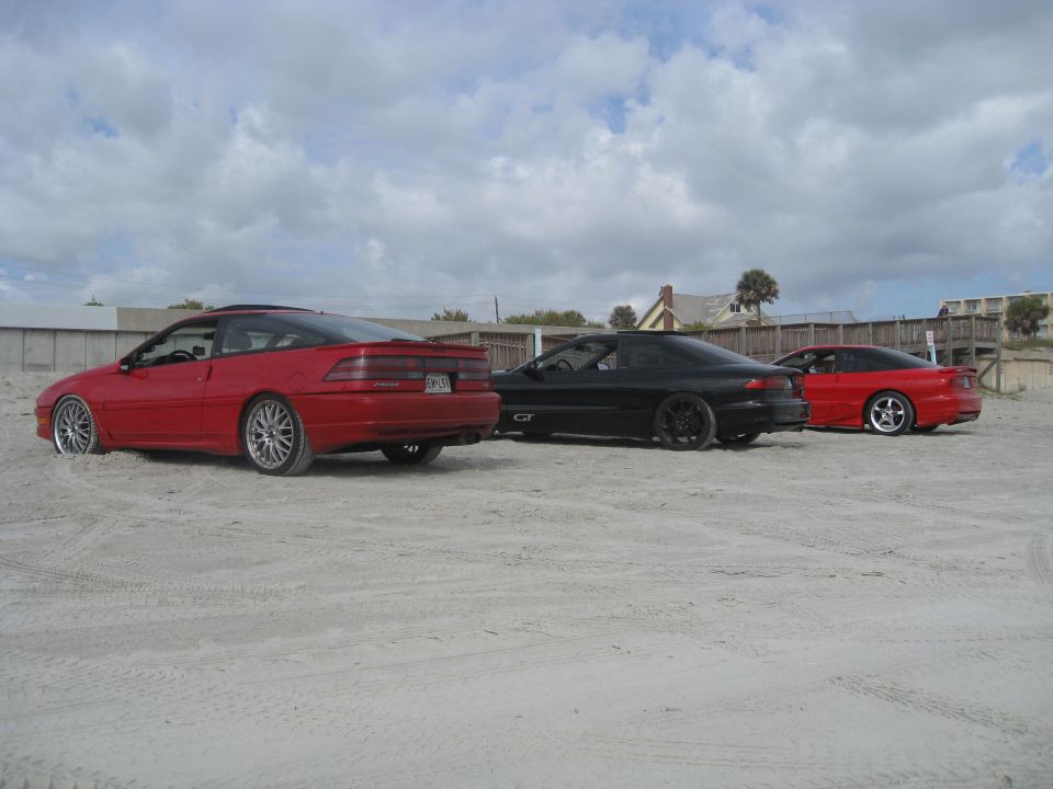 Red, black, and red coupes parked on a sandy area with buildings and sky in the background