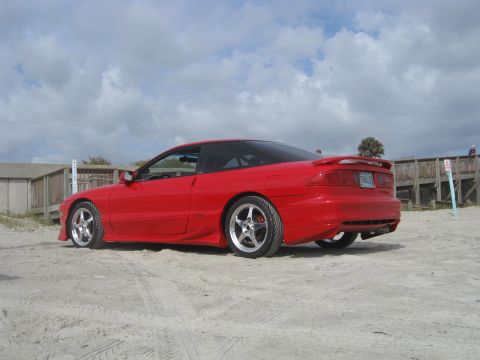 1994 Ford Probe - A red 1994 Ford Probe is parked on a sandy area with a wooden boardwalk in the background.