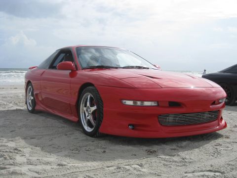 1994 Ford Probe - A red 1994 Ford Probe sits on the sand near the ocean, with another car partially visible behind it.