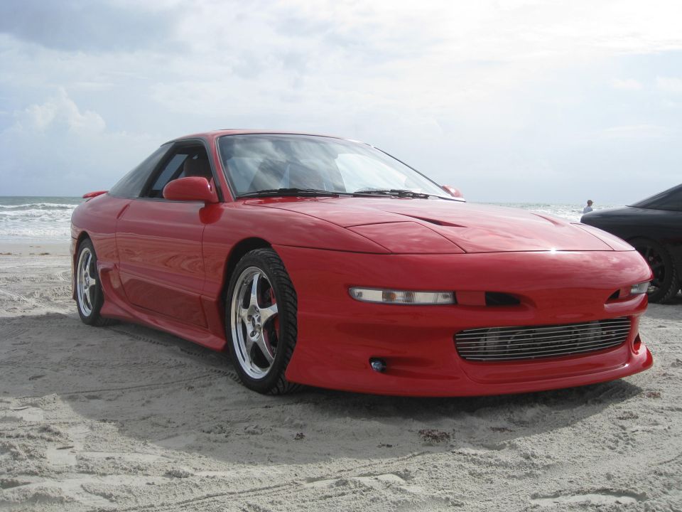Red 1994 Ford Probe coupe parked on a sandy beach with the ocean in the background