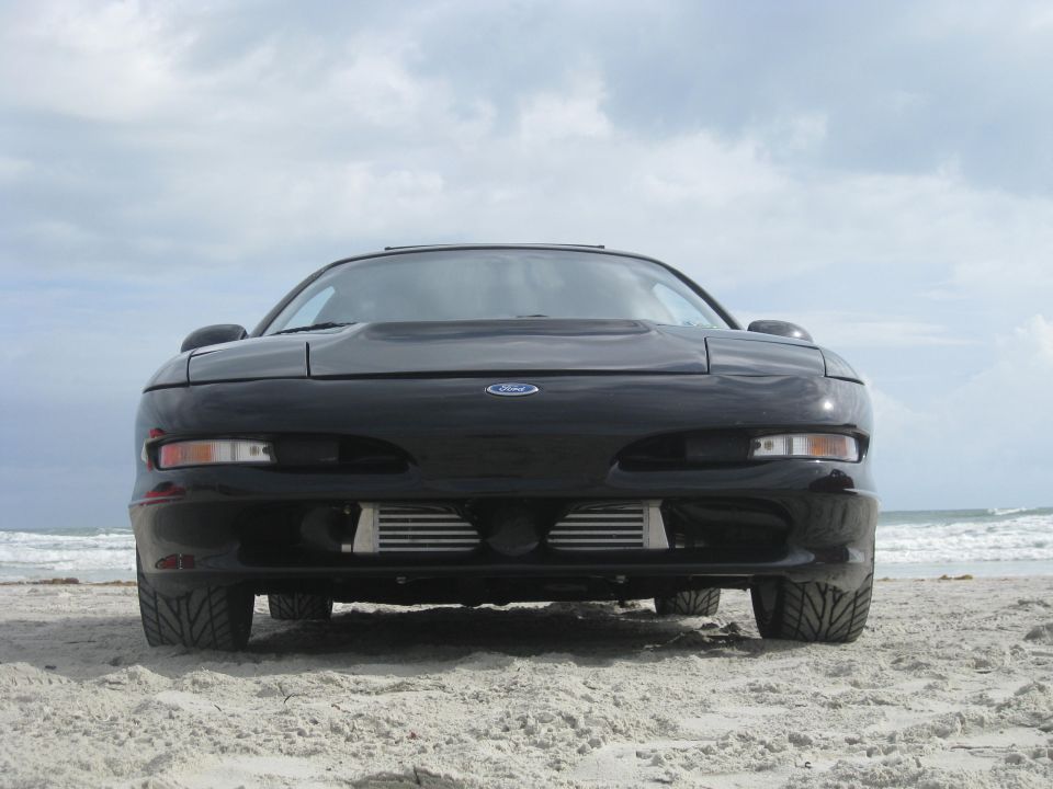 Front view of a black 1994 Ford Probe parked on a sandy beach with the ocean in the background