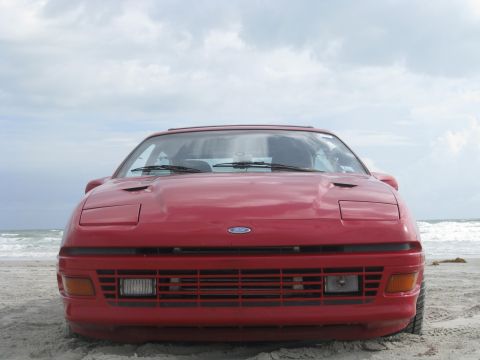 1994 Ford Probe - A red 1994 Ford Probe is parked on the sand near the ocean under a cloudy sky.