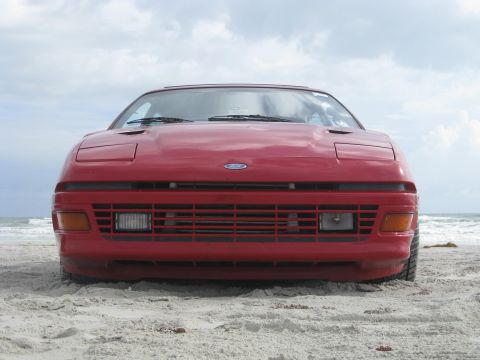 1994 Ford Probe - A red Ford Probe is parked on the sand near the ocean under a cloudy sky.