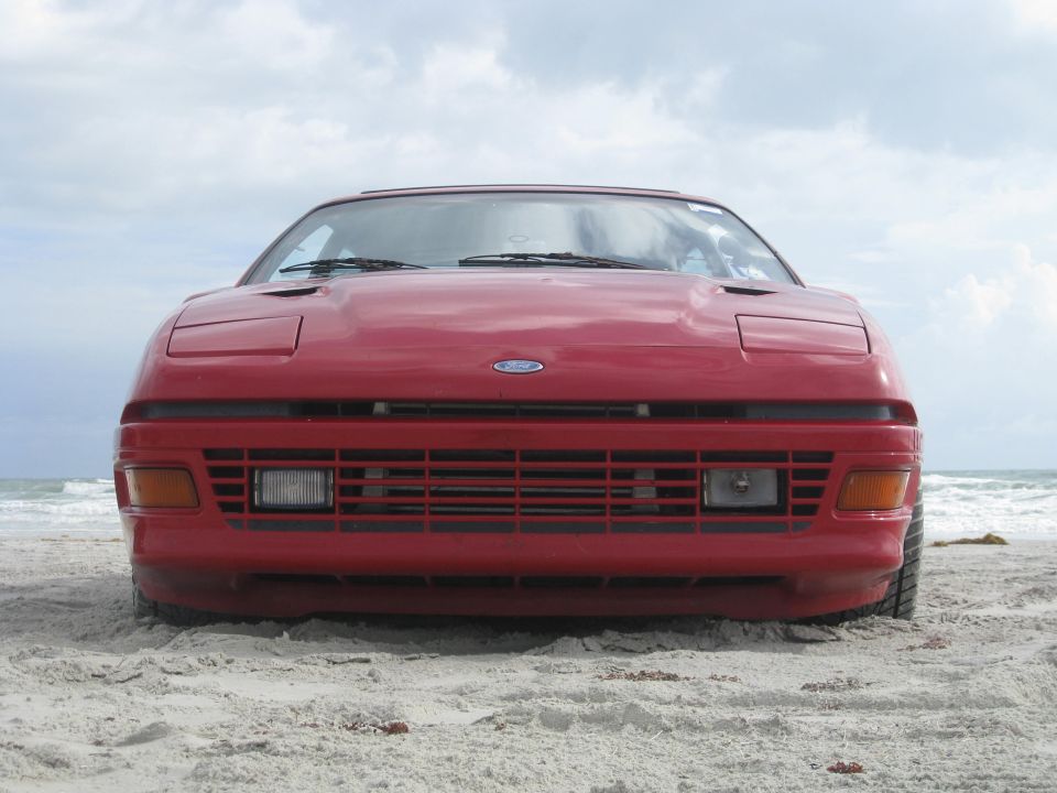 Front view of a red Ford Probe parked on a sandy beach with the ocean in the background