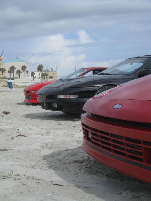 1994 Ford Probe - Several Ford Probes are lined up on the sand near the ocean under a cloudy sky.
