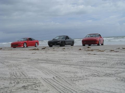 1994 Ford Probe - Three cars are parked on a sandy beach under a cloudy sky, with the ocean waves visible behind them.