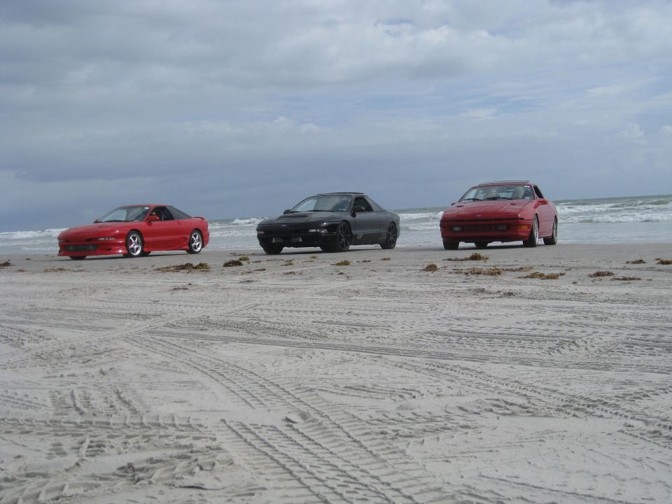 Three cars, two red and one black, parked on a sandy beach with the ocean in the background