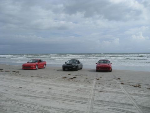 1994 Ford Probe - Three sports cars sit on the sand near the ocean under a cloudy sky.