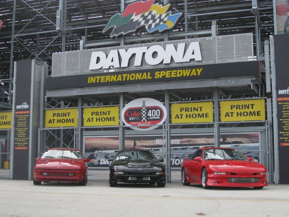 Three sports cars, two red and one black, parked in front of the Daytona International Speedway entrance