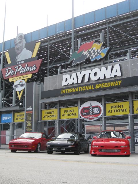 1994 Ford Probe - Three sports cars are parked outside Daytona International Speedway, with prominent signage for the venue and a Coca-Cola promotion.