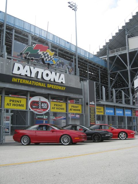 1994 Ford Probe - Three sports cars are parked outside the Daytona International Speedway, with the iconic sign visible in the background.