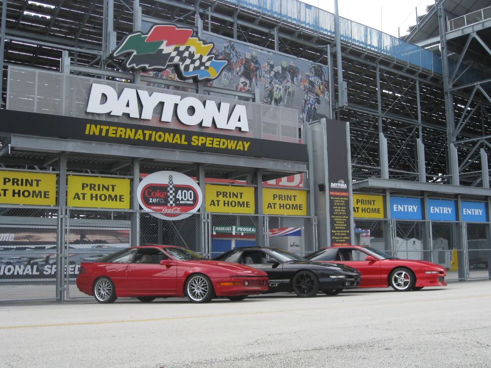 Three Ford Probes parked in front of Daytona International Speedway entrance. The red car on the left has silver wheels, the black car in the middle has black wheels, and the red car on the right has silver wheels