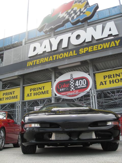 1994 Ford Probe - A black sports car is parked at the Daytona International Speedway, with a red car partially visible on the left.