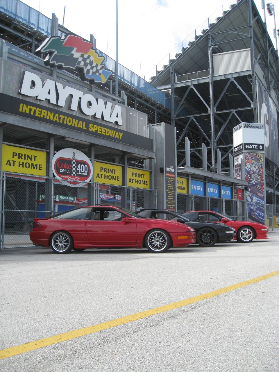 Three coupes parked in front of Daytona International Speedway entrance. The first car is red, the second is black, and the third is red