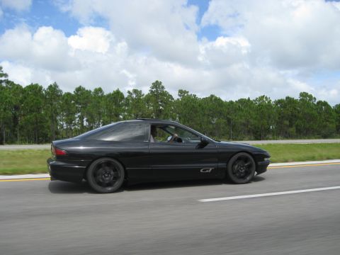 1994 Ford Probe - A black 1994 Ford Probe GT is captured in motion on a highway, with a line of trees in the background under a cloudy sky.