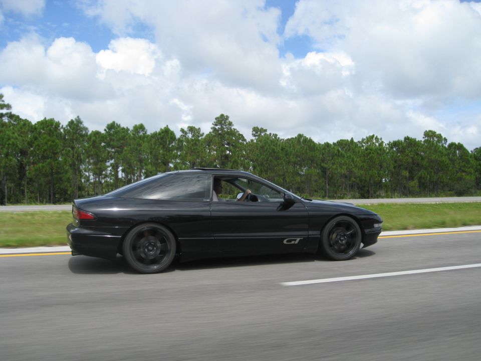 Black 1994 Ford Probe GT driving on a highway, side view. The car has black wheels and a sunroof