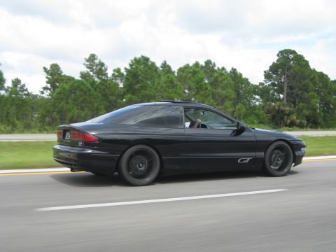 1994 Ford Probe - A black 1994 Ford Probe GT is captured in a side profile shot while driving on a highway with trees in the background.