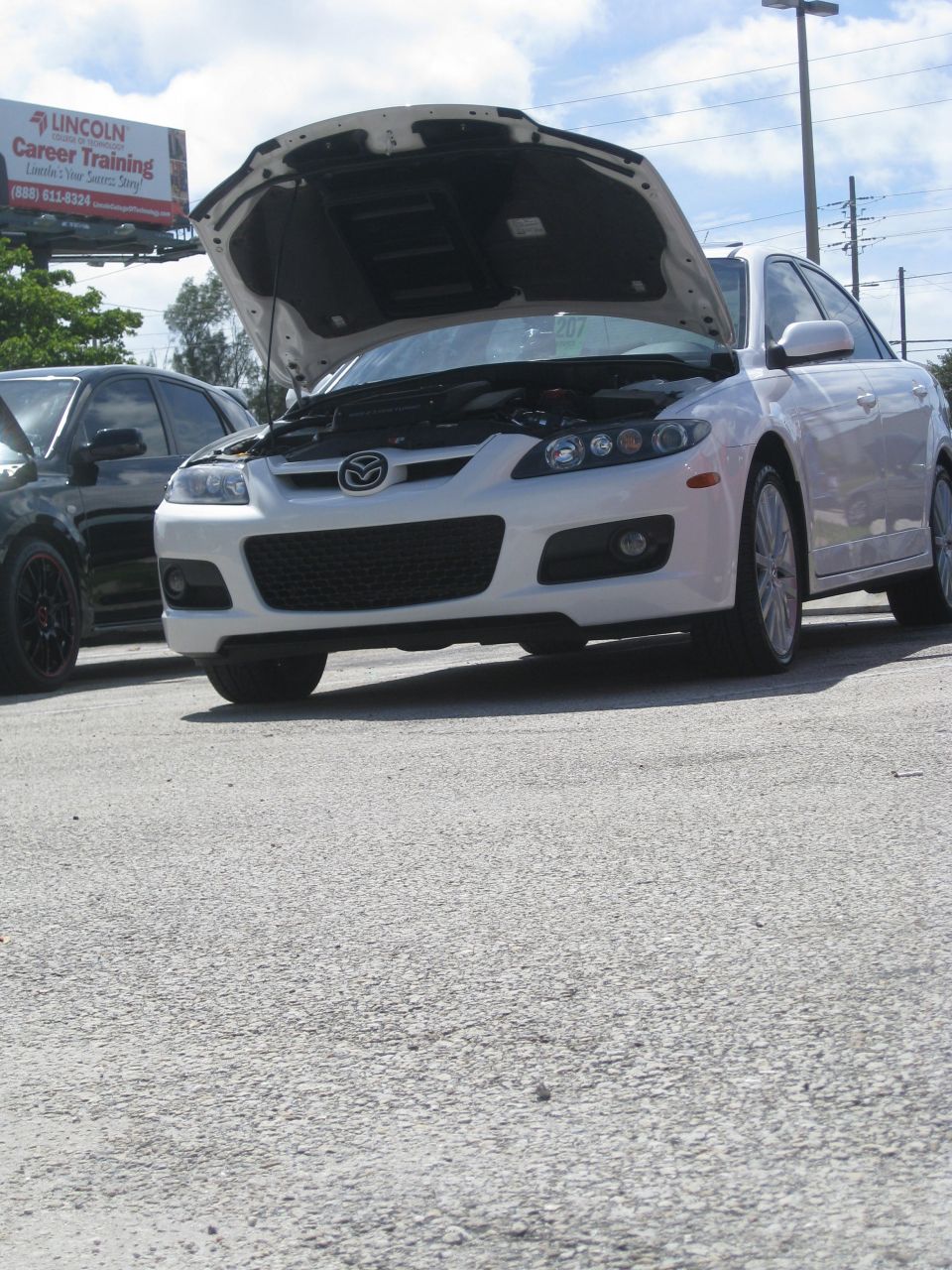 White Mazda 6 sedan with hood open, front view. Black car partially visible to the left