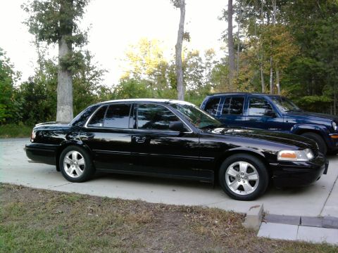 1985 Ford LTD Crown Victoria - A black full-size four-door sedan shown in profile from the passenger side, parked on a concrete driveway with silver five-spoke alloy whee…
