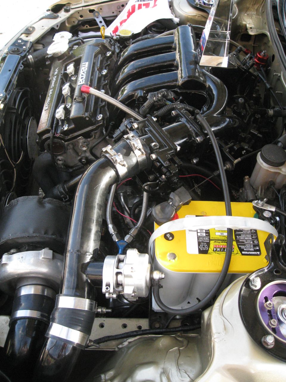 Engine bay with a turbocharger, black intake manifold, and yellow Optima battery. Some red and blue fittings are visible