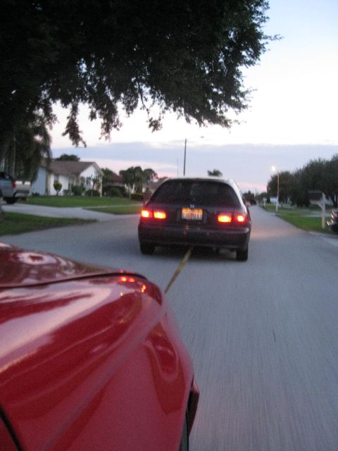 1994 Ford Probe - A red car tows a dark hatchback down a suburban street at dusk.