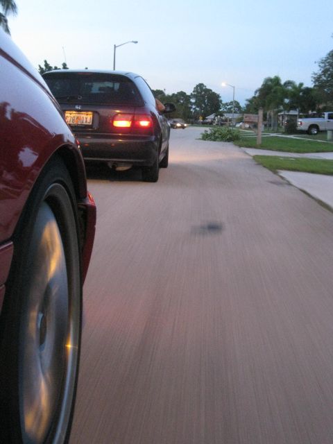 1994 Ford Probe - A red car's front wheel is visible in the foreground, while a dark hatchback drives ahead on a suburban street.