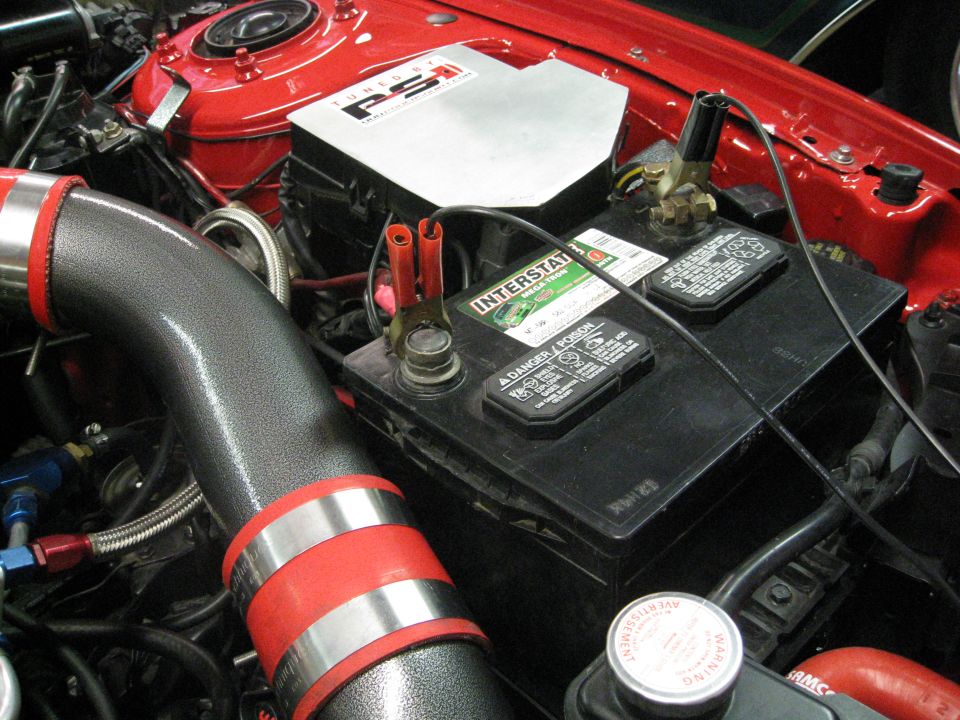 Close-up of a car's engine bay showing an Interstate battery, custom intake pipe, and red painted engine components