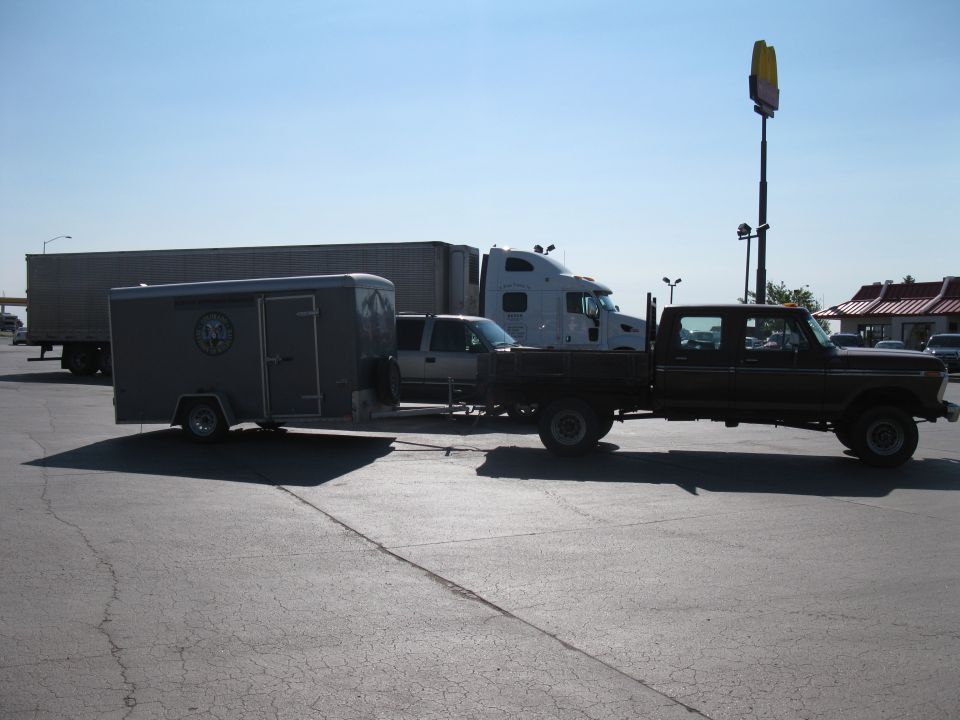 1977 Ford F-250 crew cab flatbed truck towing a grey enclosed trailer, parked next to a semi-truck