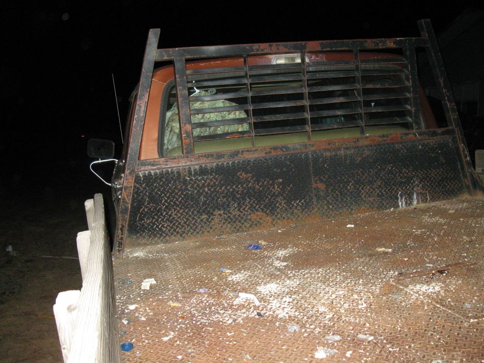 Rusty flatbed truck with a metal rack behind the cab. The cab is orange and the interior has camouflage seat covers