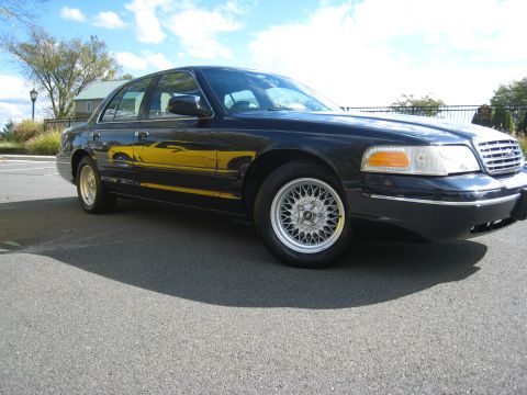 1999 Ford Crown Victoria - A dark-colored sedan is viewed from a low angle, showing its front quarter and side profile.
