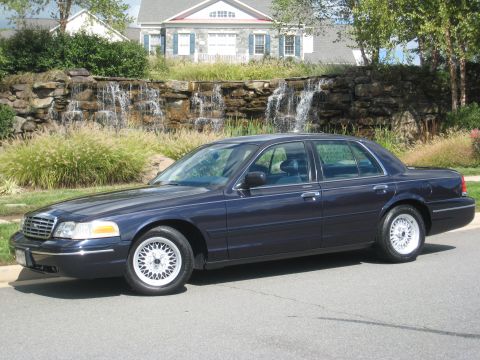 1999 Ford Crown Victoria - Dark blue sedan parked on street with a house and waterfall behind it.
