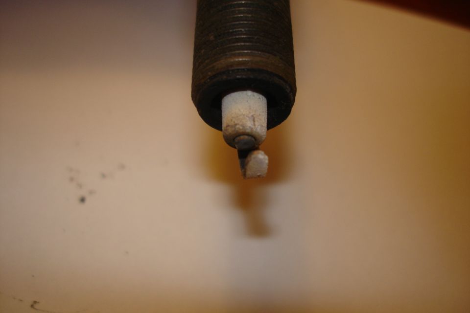 Spark plug with worn electrode and ceramic insulator against a light brown background. "SUPERMOTORS www.supermotors.net" logo in lower right