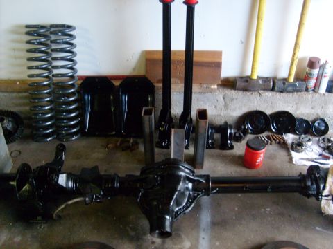 1986 Ford Bronco - Various vehicle parts, including an axle and springs, are organized on the floor, possibly for repair or upgrade.