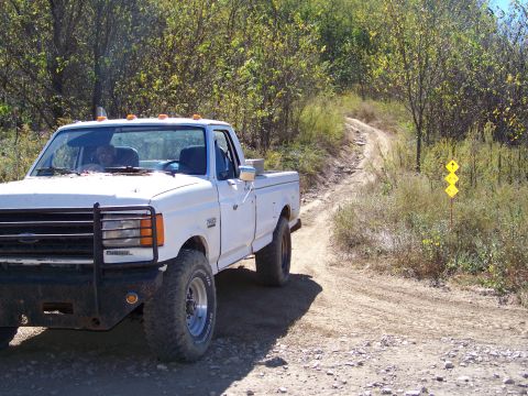 1987 Ford F-250 - White 1987 Ford F-250 pickup truck navigates a rough dirt road with trees on either side.