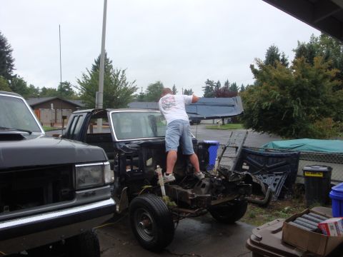 1989 Ford Bronco - A man in a white T-shirt and denim shorts leans into the stripped cab of a dismantled Ford truck in a driveway, its front clip, fenders and…