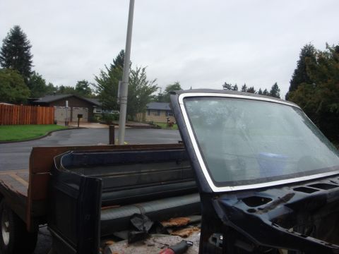 1989 Ford Bronco - A dark blue truck cab shell sits gutted in a wet suburban driveway, windshield still in its chrome trim while the cowl and firewall are str…