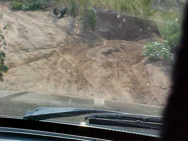 View from inside a vehicle looking out at a dirt road and hillside with brush. Windshield wipers are visible